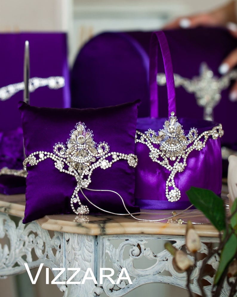 May include: A purple satin ring pillow and flower girl basket with silver rhinestone embellishments. The pillow and basket are decorated with a crown-shaped design.