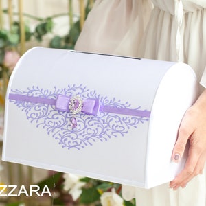 Op de afbeelding: Witte bruiloftkaartendoos met een lavendelkleurig lint en een decoratieve strik met juwelen. De doos heeft een bloemmotief in lavendel.