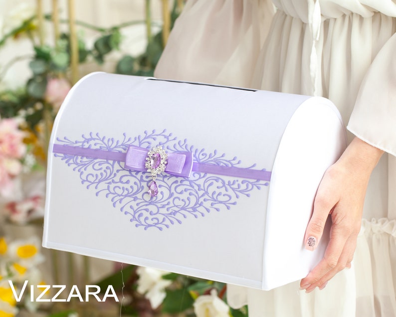 Op de afbeelding: Witte bruiloftkaartendoos met een lavendelkleurig lint en een decoratieve strik met juwelen. De doos heeft een bloemmotief in lavendel.
