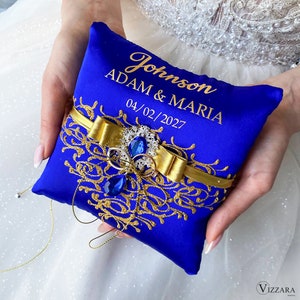 May include: Blue satin ring pillow with gold embroidery and a gold ribbon with a blue jewel accent. The pillow is personalized with the text "Johnson ADAM & MARIA 04/02/2027".