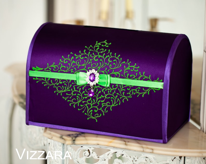 May include: A purple satin wedding card box with a green and white floral design and a purple jewel accent.
