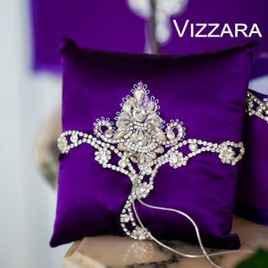 May include: A purple satin ring pillow with a silver rhinestone design and the word "VIZZARA" printed on the background.