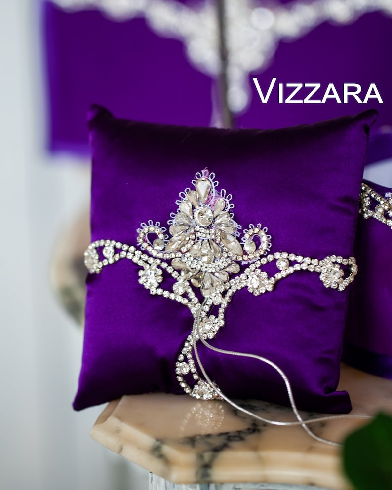 May include: A purple satin ring pillow with a silver rhinestone design and the word "VIZZARA" printed on the background.