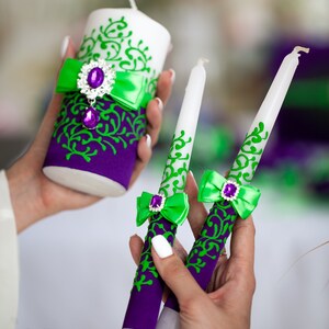 May include: Three white candles with green and purple decorative accents. The candles are decorated with a green ribbon and a purple jewel. The candles are for a wedding ceremony.