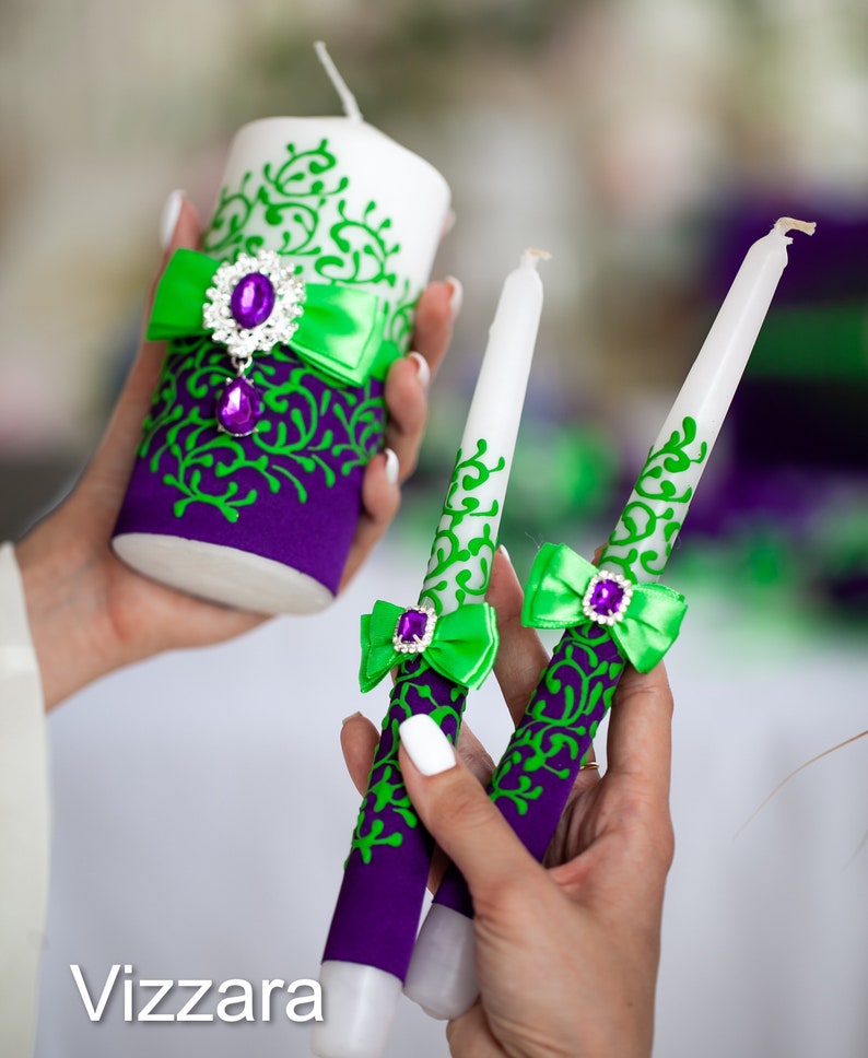 May include: Three white candles with green and purple decorative accents. The candles are decorated with a green ribbon and a purple jewel. The candles are for a wedding ceremony.