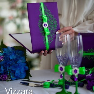 May include: A purple and green wedding guest book with a green ribbon and a jeweled bow. Two clear glass champagne flutes with green and purple embellishments are also pictured.