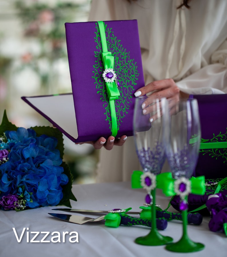 May include: A purple and green wedding guest book with a green ribbon and a jeweled bow. Two clear glass champagne flutes with green and purple embellishments are also pictured.