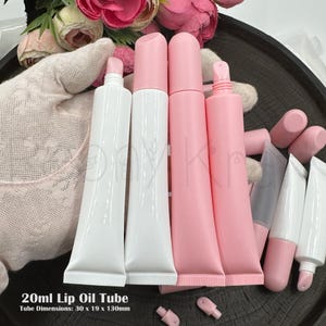 May include: A collection of 20ml lip oil tubes measuring 30 x 19 x 130mm. The set includes glossy white tubes with pink caps and matte pink tubes with matching pink caps. Some tubes are shown with caps removed, revealing the applicator tips.