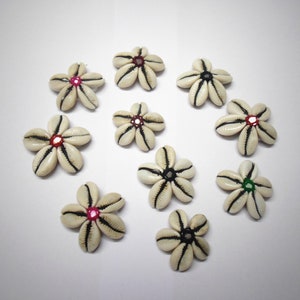 Op de afbeelding: Tien witte schelpvormige ornamenten in de vorm van een bloem met een zwarte rand en gekleurde centra. De centra zijn rood, roze, groen en zwart.