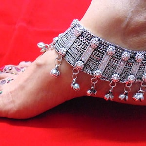 Può includere: Cavigliera d'argento con un designo decorativo e campanelle pendenti. La cavigliera è indossata su un piede con disegni all'henné e smalto rosso.