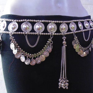 May include: A silver belly dance belt with a row of round, decorative medallions and dangling chains with coin-like charms. The belt has a tassel hanging from the center.