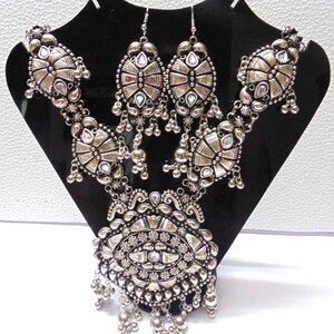 May include: A silver necklace and earrings set with intricate designs and clear stones. The necklace features a large pendant with a central eye-like design and multiple layers of dangling beads.