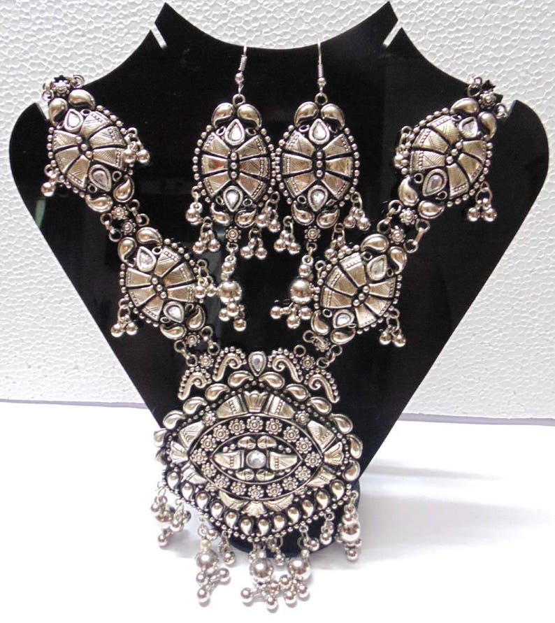 May include: A silver necklace and earrings set with intricate designs and clear stones. The necklace features a large pendant with a central eye-like design and multiple layers of dangling beads.