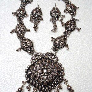 May include: A silver necklace and earrings set with intricate, ornate designs. The necklace features a large pendant with a central stone and dangling beads. The earrings are similarly detailed with dangling beads.