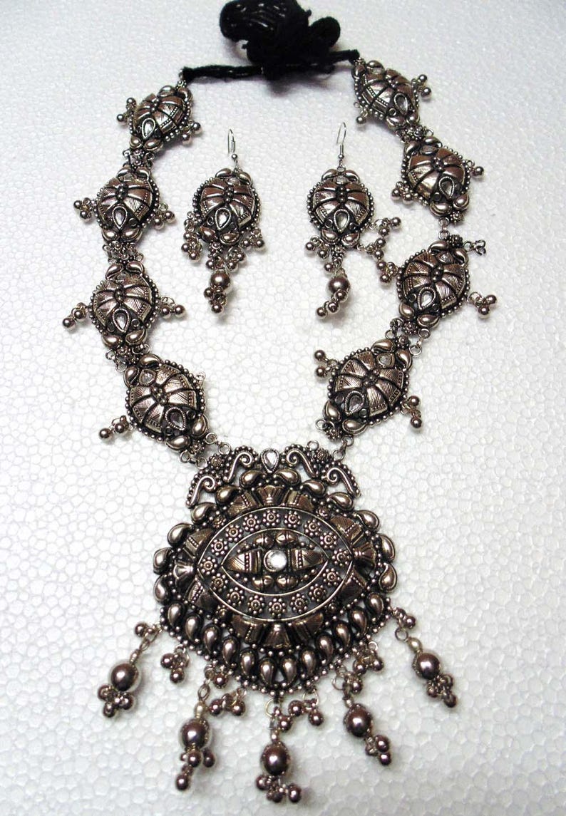May include: A silver necklace and earrings set with intricate, ornate designs. The necklace features a large pendant with a central stone and dangling beads. The earrings are similarly detailed with dangling beads.