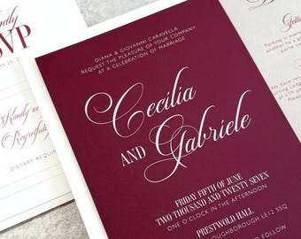 May include: Burgundy wedding invitation with white script text. The invitation reads "Cecilia and Gabriele" and includes event details: date, time, and location. Additional cards include RSVP and detail information. A rose gold paper clip is in the foreground.