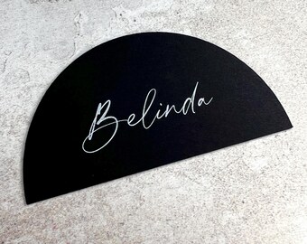 May include: A black, semi-circular place card with the name "Belinda" in elegant white script. The card is set against a textured, light gray background, creating a contrast that highlights the name.