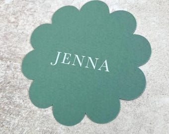 May include: A sage green, scalloped-edge place card with the name "JENNA" printed in white. The card is circular with a floral-like shape. The background is a light gray, textured surface.