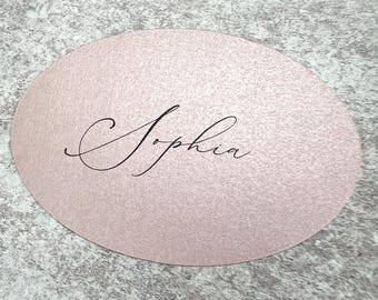 May include: A pale pink, oval-shaped place card with the name "Sophia" written in elegant black script. The card has a textured surface and is set against a gray, speckled background.