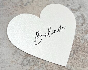 May include: A heart-shaped place card with the name "Belinda" written in elegant black script. The card is a soft white color with a subtle textured finish. Ideal for weddings or special events.