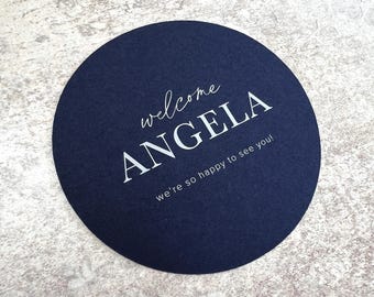 May include: A navy blue circular coaster with the text "welcome ANGELA we're so happy to see you!" in white. The coaster is made of a textured material and is likely used for drinks.