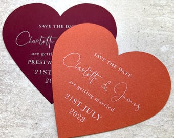 May include: Two heart-shaped wedding announcements. One is a deep burgundy color, the other is a burnt orange. Both have white script that reads "Save the Date" and the names "Charlotte" and "James" with the wedding date.