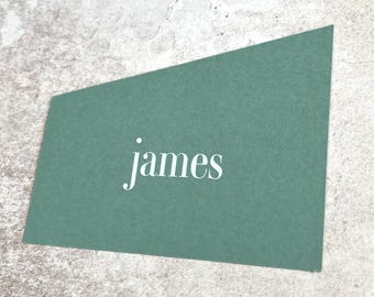 May include: A teal-colored business card with the name "james" in white script. The card has a unique angled shape and is set against a textured, light gray surface. The design is simple and elegant.