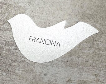 May include: A silver-colored dove-shaped decorative item with the name "FRANCINA" printed in black. The dove has a textured surface and is set against a gray background.