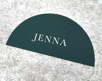 May include: A semi-circular, dark green place card with the name "JENNA" printed in white, centered on the card. The card is set against a textured, light gray surface.