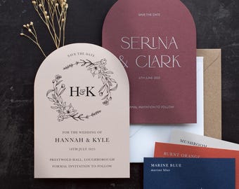May include: Arch-shaped wedding stationery set featuring a light pink card with a floral wreath and H&K monogram, a dark maroon card with Serina & Clark text, and smaller rectangular color swatch cards labeled Mushroom, Burnt Orange, and Marine Blue.
