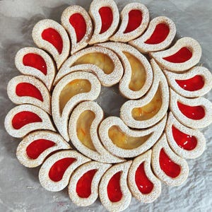 Può includere: Una disposizione circolare di pasticceria con un motivo paisley ripetuto. I pasticcini sono ripieni di gelatina rossa e gialla, spolverati di zucchero a velo e disposti a forma di ghirlanda. I prodotti da forno sono su un foglio di carta forno.
