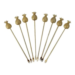 May include: A set of eight gold-toned metal cocktail picks, each featuring a textured pineapple-shaped topper at the handle end and a two-pronged fork at the base. The picks are arranged in a fan shape, showcasing the uniform metallic finish.