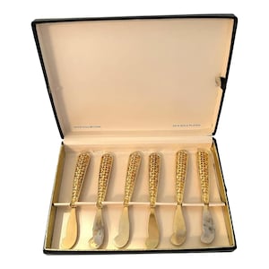 May include: A set of six gold-plated butter knives in a black box. The knives have textured handles and curved blades. The box is labeled "JANIS COLLECTION" and "24 K GOLD PLATED".