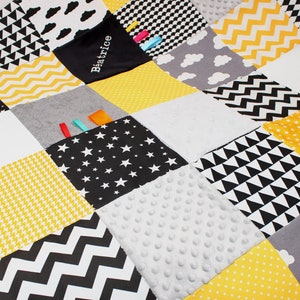 Puede incluir: Una alfombra de juego para bebés en blanco y negro con una variedad de texturas y patrones. La alfombra está hecha de diferentes telas, incluyendo una tela suave y peluda, una tela texturizada y una tela lisa. La alfombra también tiene una variedad de colores, incluyendo negro, blanco, amarillo y gris. La alfombra tiene una etiqueta con el nombre "Beatrice" en ella.