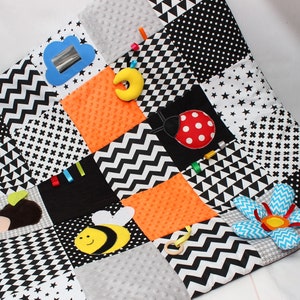 May include: A black and white patchwork baby play mat with various textures and shapes. Features include a cloud with a mirror, a yellow crescent moon, an orange square, a ladybug, a bee, and a flower. The mat is designed with different patterns.