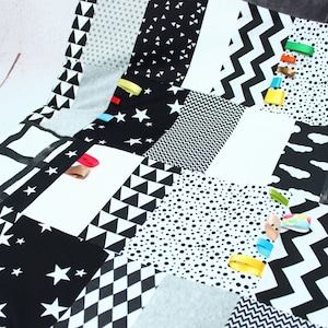 May include: Black and white baby activity play mat with various textures and patterns, including stars, triangles, polka dots, and zigzags. The mat has colorful ribbons attached to the edges.