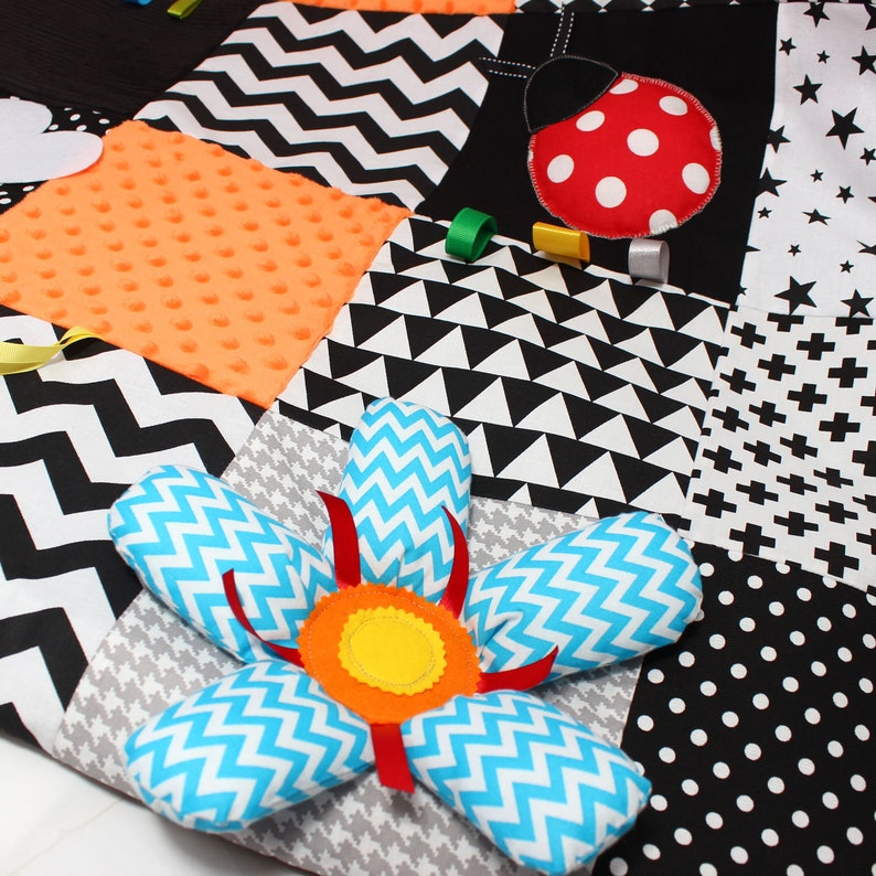 Puede incluir: Una colorida alfombra de juego para beb&eacute;s de patchwork con varias texturas y patrones. Incluye una flor grande con p&eacute;talos de chevron azules y blancos, una mariquita y formas geom&eacute;tricas en negro, blanco y naranja. Incluye peque&ntilde;os anillos de colores.