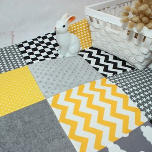May include: A patchwork play mat with various patterns, including stars, polka dots, and zigzags in yellow, gray, and black. A white ceramic bunny sits on the mat near a white wicker basket.
