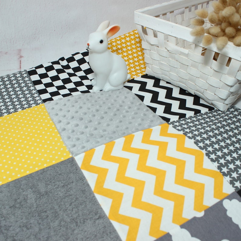 May include: A patchwork play mat with various patterns, including stars, polka dots, and zigzags in yellow, gray, and black. A white ceramic bunny sits on the mat near a white wicker basket.