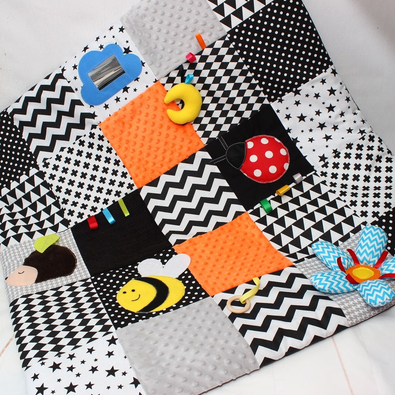 Puede incluir: Manta de juego para beb&eacute;s con dise&ntilde;o de patchwork en blanco y negro. Incluye una nube con espejo, una luna amarilla, cuadrados naranjas, una mariquita, una abeja y una flor. La manta presenta diversas texturas y patrones para el juego sensorial.