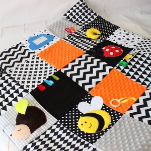 Puede incluir: Una alfombra de juego para beb&eacute;s con dise&ntilde;o de patchwork en negro, blanco, gris y naranja. Incluye una mariquita, una abeja, un erizo, una nube y cintas de colores. Incluye un anillo de madera y una media luna amarilla.