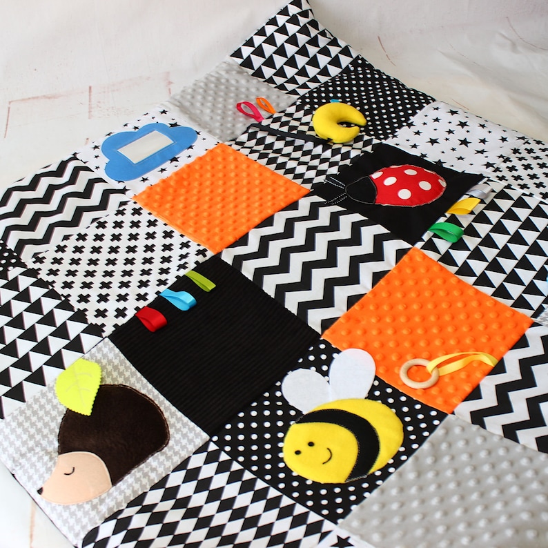 Puede incluir: Una alfombra de juego para beb&eacute;s con dise&ntilde;o de patchwork en negro, blanco, gris y naranja. Incluye una mariquita, una abeja, un erizo, una nube y cintas de colores. Incluye un anillo de madera y una media luna amarilla.