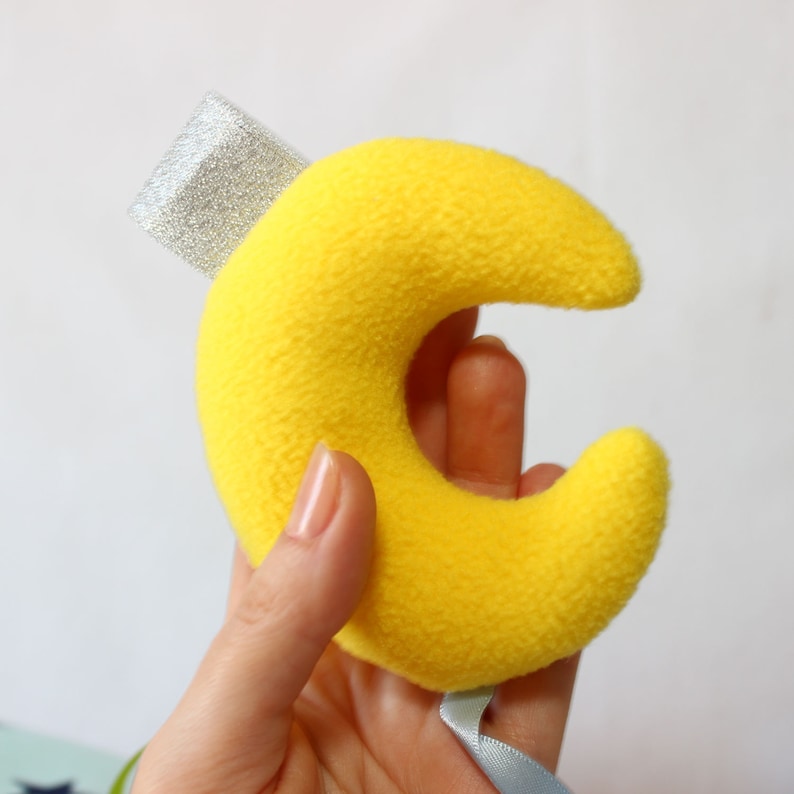 Puede incluir: Una luna creciente amarilla de tela de felpa, con un detalle de purpurina plateada y una cinta azul claro. La luna es un objeto suave y texturizado, posiblemente para una habitaci&oacute;n infantil o de beb&eacute;.