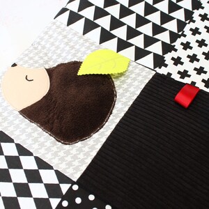 Puede incluir: Manta de actividades para beb&eacute;s con un dise&ntilde;o de erizo y una hoja amarilla. La manta presenta varios patrones geom&eacute;tricos en blanco, negro y gris, con cintas rojas y azules. El erizo tiene un cuerpo marr&oacute;n y una cara beige.