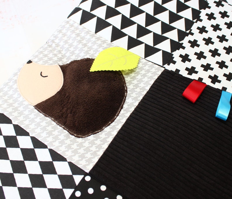 Puede incluir: Manta de actividades para beb&eacute;s con un dise&ntilde;o de erizo y una hoja amarilla. La manta presenta varios patrones geom&eacute;tricos en blanco, negro y gris, con cintas rojas y azules. El erizo tiene un cuerpo marr&oacute;n y una cara beige.
