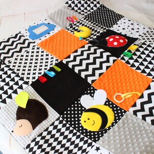 Puede incluir: Un tapete de juego para beb&eacute;s de patchwork en blanco, negro y naranja. El tapete presenta una variedad de texturas y formas, incluyendo una nube, una media luna, una mariquita, una abeja y un erizo. El tapete tiene varios patrones, incluyendo lunares, chevrones y diamantes.