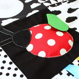 Puede incluir: Alfombra de juego para beb&eacute;s en blanco y negro con un dise&ntilde;o de mariquita roja. La mariquita tiene lunares blancos y una hoja verde. La alfombra presenta varios patrones, incluyendo nubes y lunares, y cintas de colores.