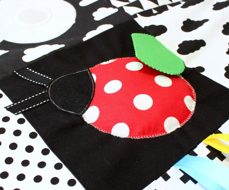 Puede incluir: Alfombra de juego para beb&eacute;s en blanco y negro con un dise&ntilde;o de mariquita roja. La mariquita tiene lunares blancos y una hoja verde. La alfombra presenta varios patrones, incluyendo nubes y lunares, y cintas de colores.