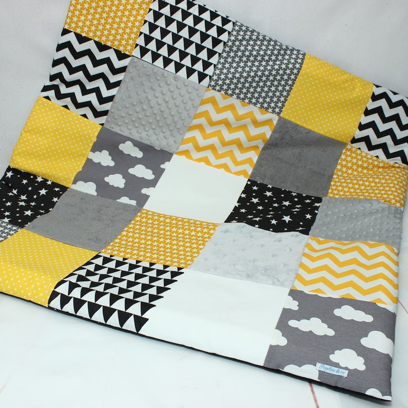 May include: A patchwork quilt featuring a variety of patterns and colors. The quilt is composed of squares in yellow, black, white, and gray. Patterns include stars, chevrons, triangles, and clouds. The quilt has a soft, textured appearance.