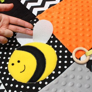 Puede incluir: Una colorida manta sensorial para beb&eacute;s con un aplique de abeja amarilla y negra sonriente. La manta presenta varias texturas y patrones, incluyendo lunares, un dise&ntilde;o en zigzag y un anillo de madera con una cinta amarilla.